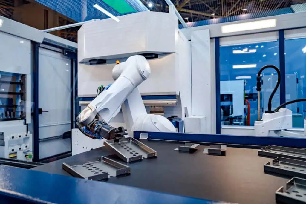 A robotic arm in a modern factory assembles metal components on a worktable, surrounded by advanced machinery and blue-tinted glass panels.