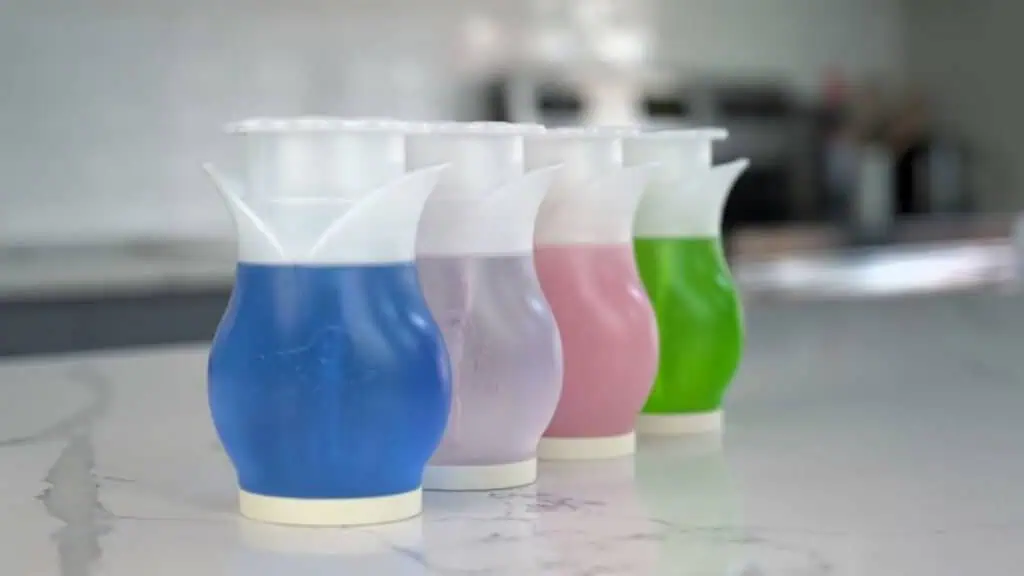 Four flower-shaped automatic toilet bowl cleaners in blue, clear, pink, and green, are lined up on a white marble countertop in a modern kitchen setting.
