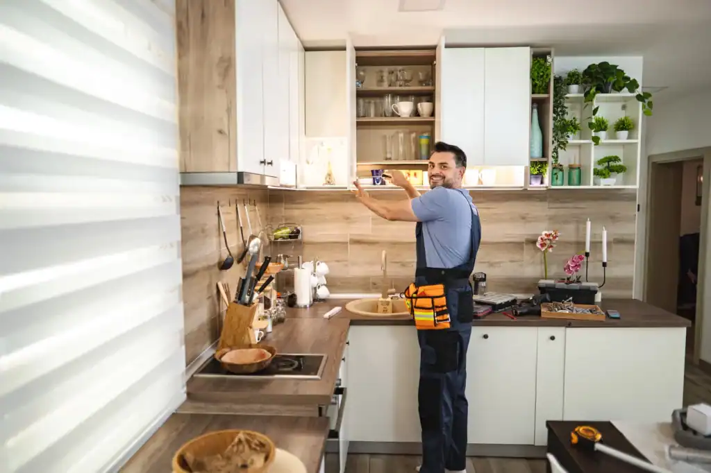A man wearing a tool belt is fixing or installing a kitchen cabinet, holding a power drill and smiling at the camera. The kitchen is modern with white cabinets, plants, and wooden accents.