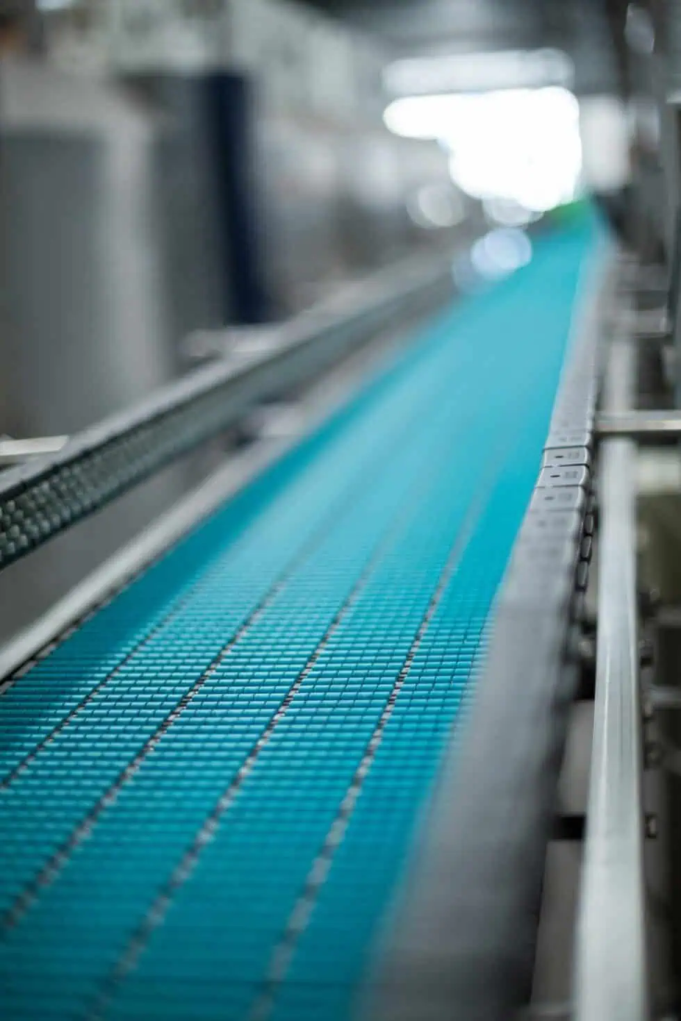 A close-up view of an empty blue conveyor belt in a factory or industrial setting, with the background blurred and the belt leading into the distance.