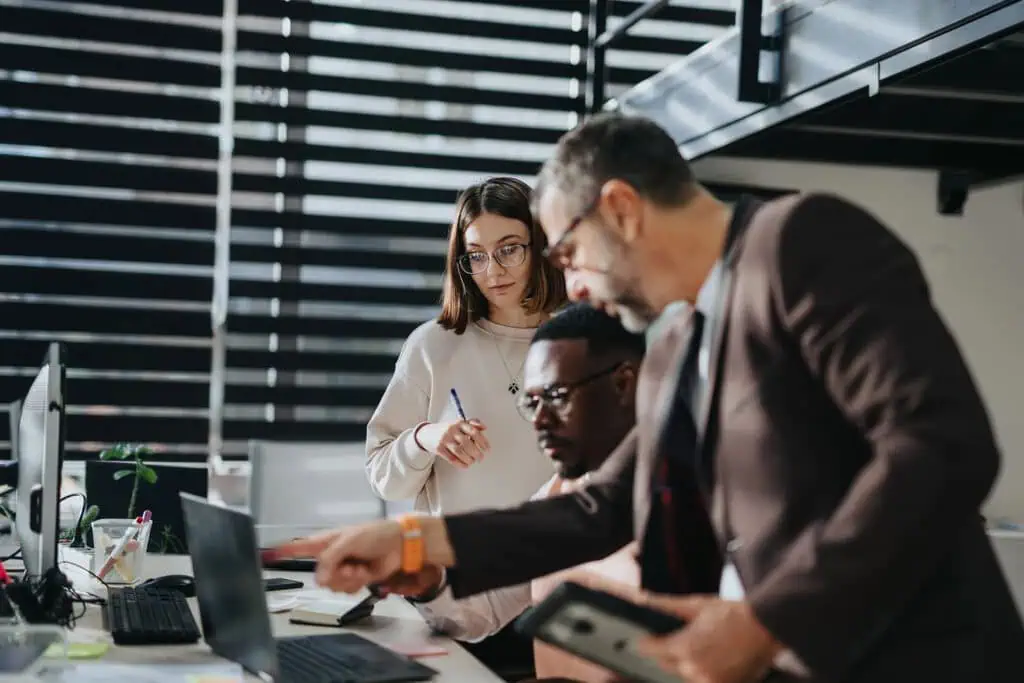 Three people in an office work together at a desk with laptops. One person points at the screen, another sits and looks at the laptop, while the third stands holding a pen and watches. The atmosphere appears collaborative.