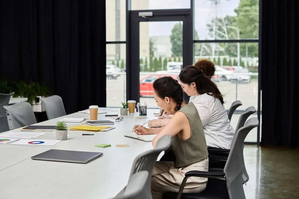 Two women sit at a conference table with notebooks and laptops, working in a modern office space with large windows and outdoor views. Charts, coffee cups, and office supplies are on the table.
