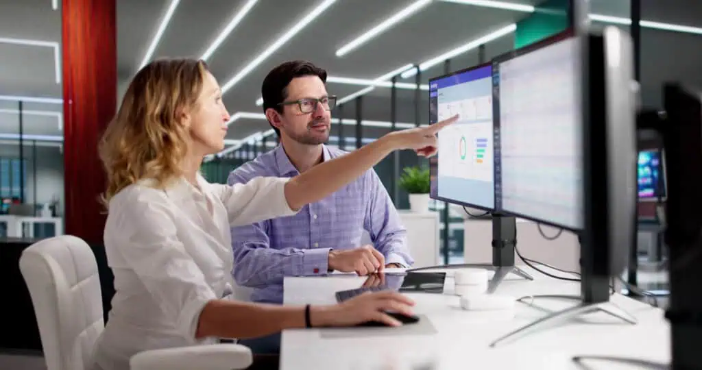 A woman and a man sit at a desk in a modern office, looking at data on dual computer monitors. The woman is pointing at the screen while the man watches attentively. Bright lights and office decor are visible in the background.