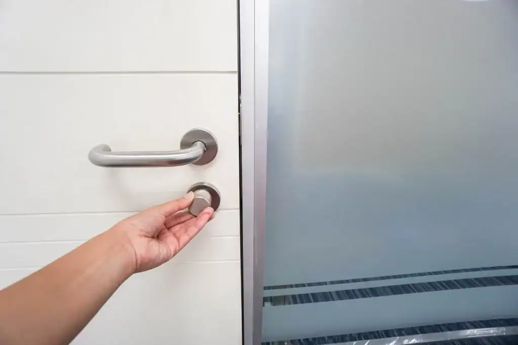 A hand is turning a round door lock on a white door next to a frosted glass partition.