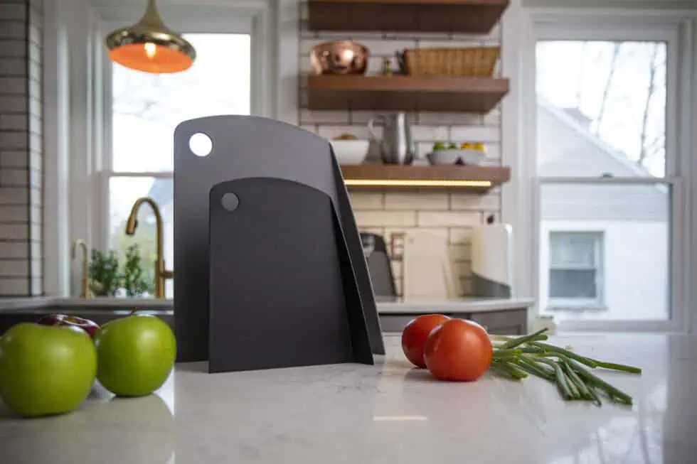 Two black cutting boards stand upright on a kitchen counter, surrounded by green apples, tomatoes, and green onions. The modern kitchen has white cabinets, large windows, and wooden shelves in the background.