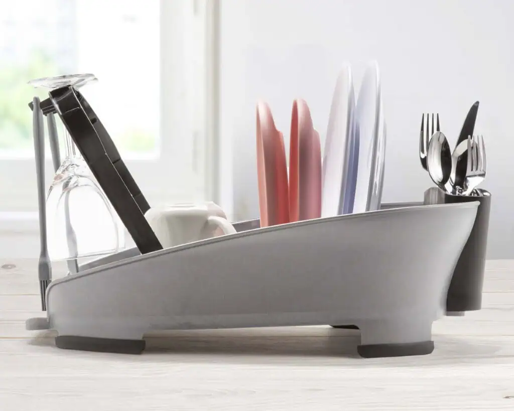A gray dish rack holding plates, bowls, glasses, utensils, and a mug sits on a light-colored kitchen counter in front of a window with soft daylight coming through.