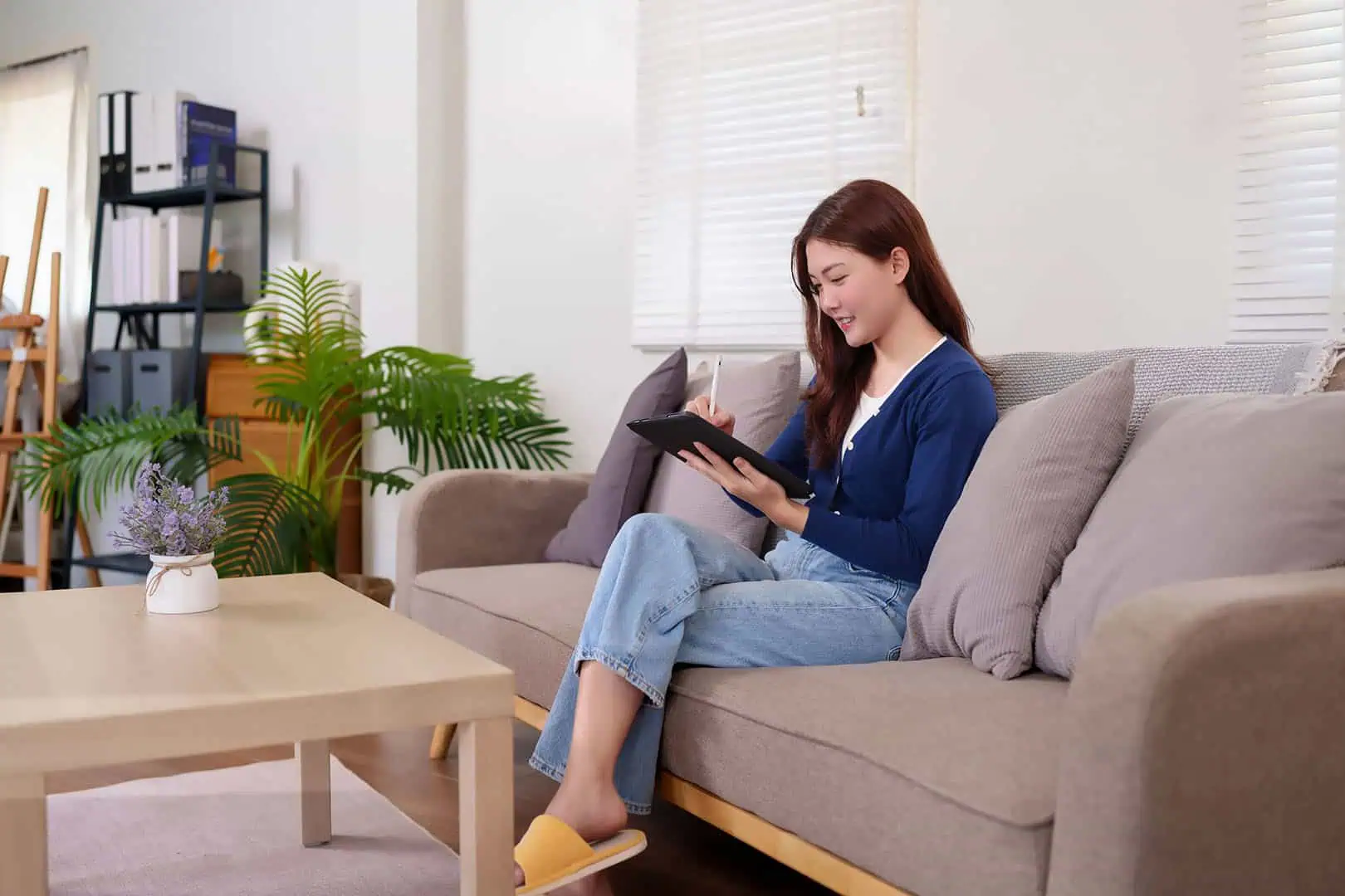 A woman sits on a sofa in a cozy living room, smiling while using a digital tablet. She wears a blue cardigan, jeans, and yellow slippers. A coffee table with a flower vase is in front of her, and plants are in the background.